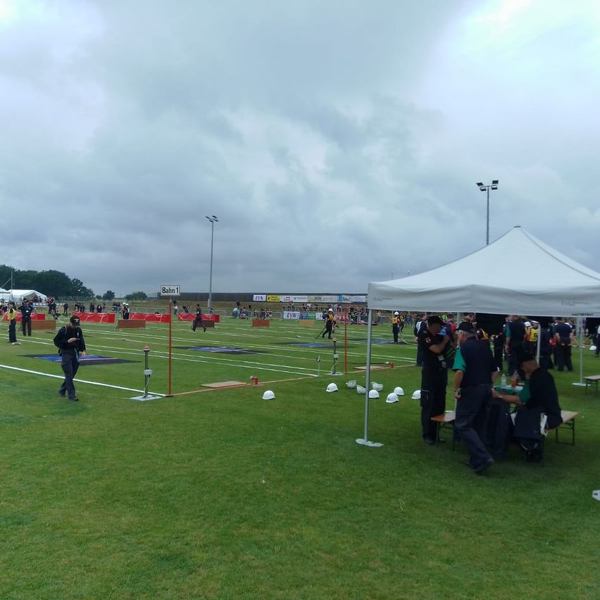 Eine Sportveranstaltung auf einem Feld mit Teilnehmern und Zuschauern unter einem weißen Zelt. Viele Menschen sind versammelt, einige tragen Hüte und Schuhe. Ein paar sitzen auf Bänken, andere stehen.