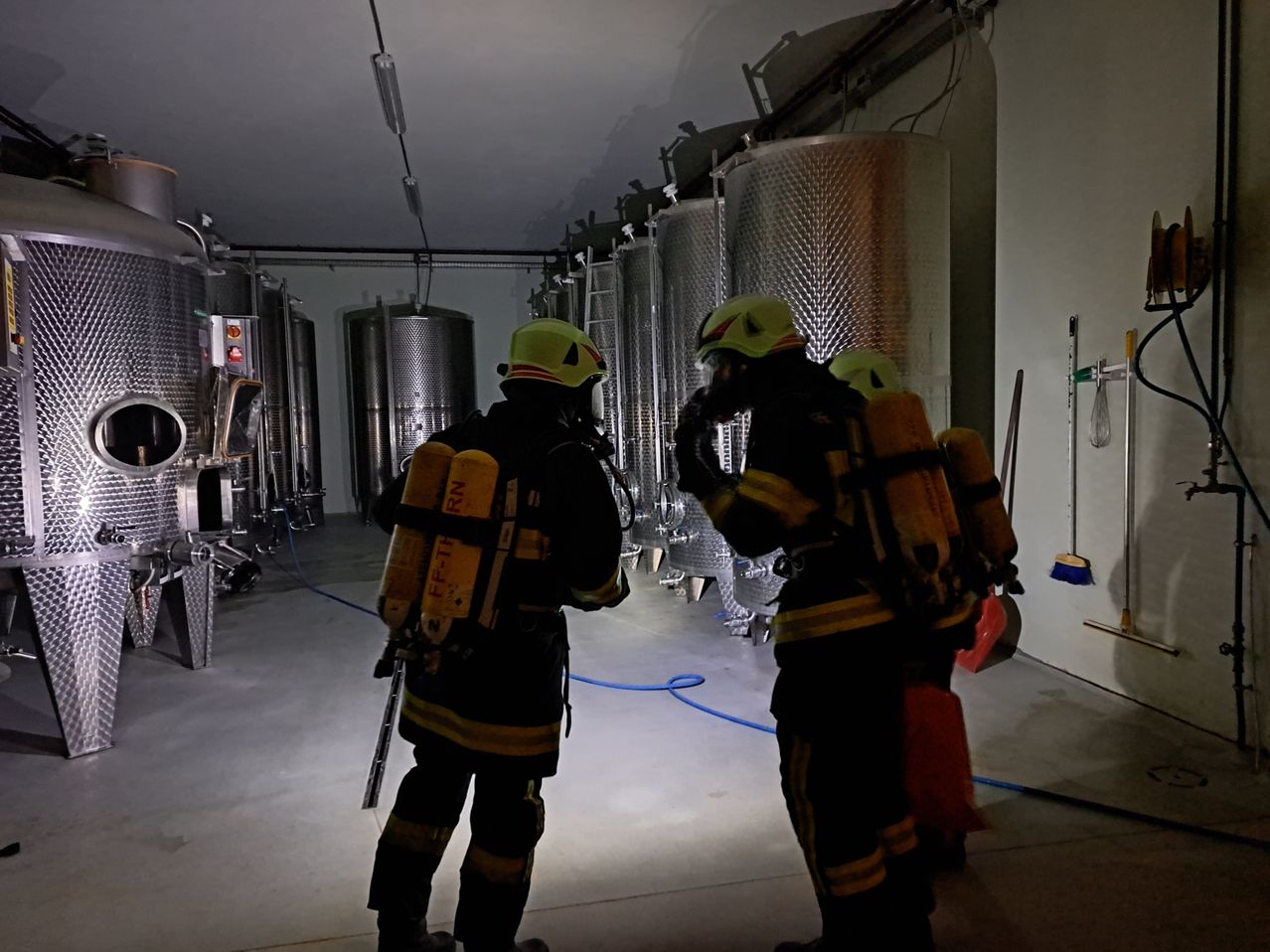 Zwei Feuerwehrleute in Schutzkleidung stehen in einem Raum mit großen Edelstahltanks. Sie stehen sich gegenüber und scheinen zu sprechen. Der Raum ist schwach beleuchtet.