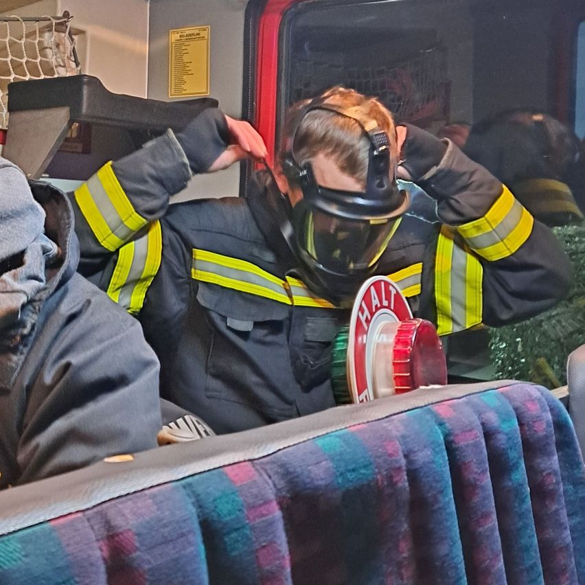 Ein Feuerwehrmann in Schutzausrüstung sitzt auf einem Bus und justiert seine Maske. Das Innere des Busses ist zu sehen, mit einem roten und weißen Schild auf dem Sitz, das 'HALT' steht.