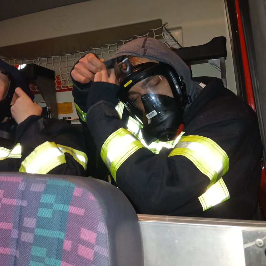 Ein Feuerwehrmann mit Gasmaske sitzt auf einer Bank in einem Fahrzeug, wahrscheinlich einem Feuerwehrwagen. Ein weiterer Feuerwehrmann steht in der Nähe. Der Innenraum ist gut beleuchtet, und es gibt reflektierende gelbe Streifen auf ihren Uniformen.