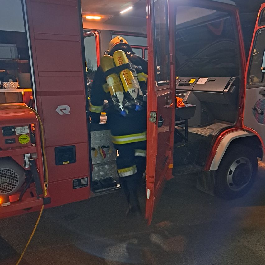 Ein Feuerwehrmann verlässt einen roten Feuerwehrwagen, trägt einen gelben Helm und eine Feuerlöschflasche. Die Tür des Wagens ist geöffnet und zeigt den Innenraum.