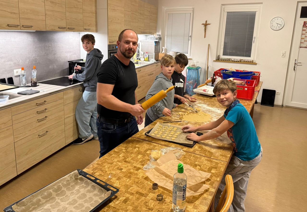 Ein Mann und vier Jungen sind in einer Küche und bereiten sich darauf vor, Kekse zu backen. Der Mann hält einen Nudelholz, und die Jungen arbeiten an Teig. Tabletts, Ablagen und eine Flasche sind auf dem Tisch. Ein Fenster mit Jalousien ist hinter ihnen.