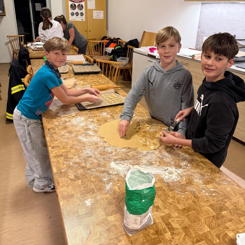 Drei Jungen arbeiten gemeinsam an einem Küchentisch und bereiten Teig mit Ausstechformen zu. Ein Junge benutzt ein Nudelholz, während die anderen beiden ihre Hände im Teig haben. Dahinter sind weitere Tische und Stühle aufgebaut, und zwei Frauen arbeiten.