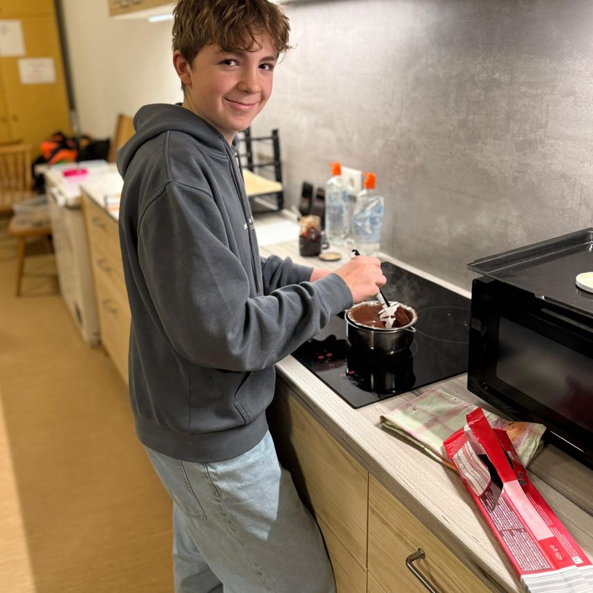 Ein junger Junge in einer grauen Kapuzenjacke rührt einen Topf auf dem Herd in einer Küche. Flaschen und andere Gegenstände stehen auf der Arbeitsplatte.