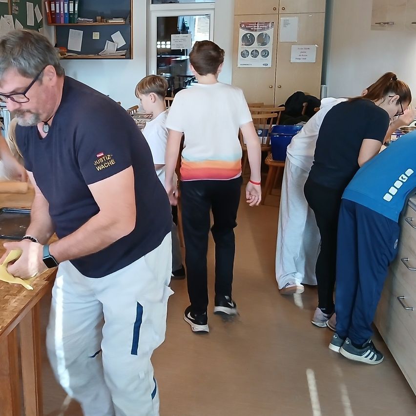 Ein älterer Mann in einem blauen T-Shirt und weißen Hosen schält eine Banane auf einem Tisch. Hinter ihm betrachten zwei Kinder und eine Frau in einem weißen Kittel etwas auf dem Boden.
