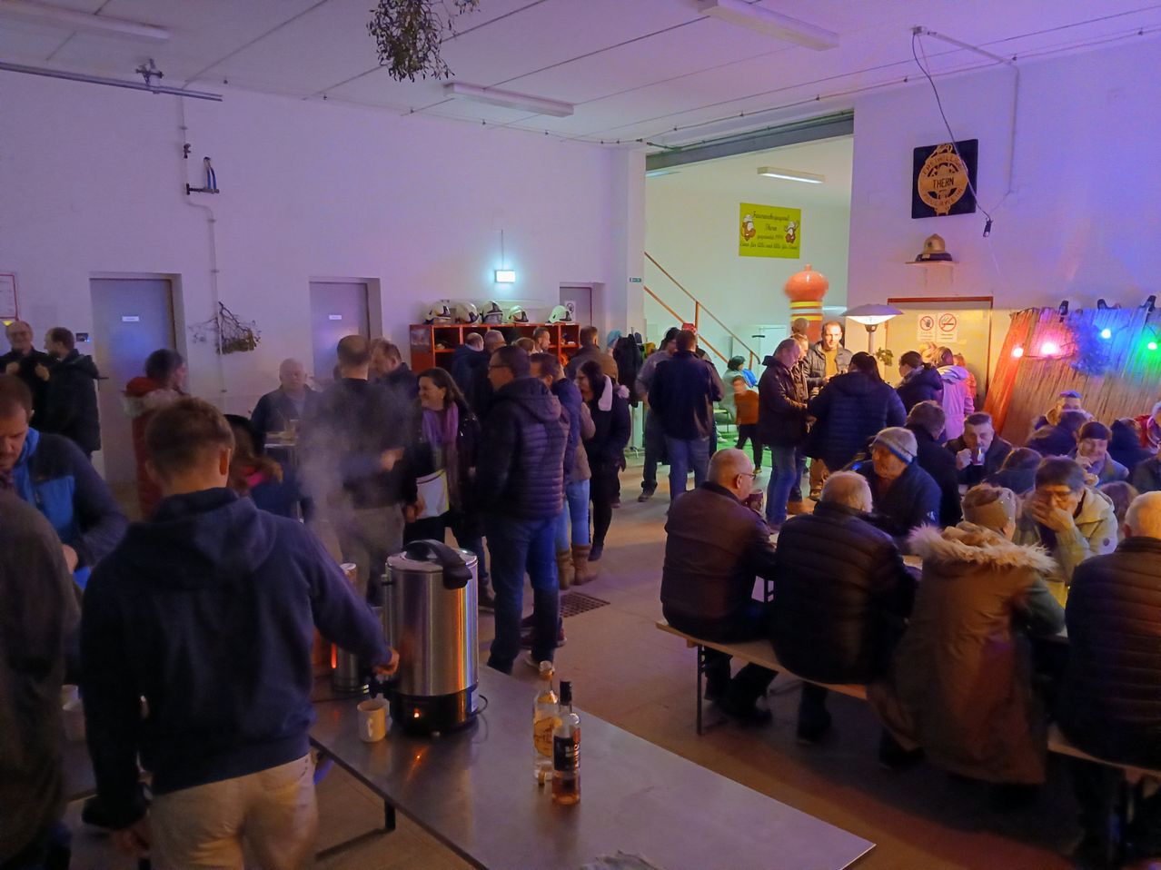 Eine Gruppe von Menschen versammelt sich in einem Saal mit Tischen und Bänken. Einige sitzen, andere stehen. Es gibt Kaffeekannen und Flaschen auf den Tischen. Der Raum ist schwach beleuchtet mit violetter Beleuchtung.