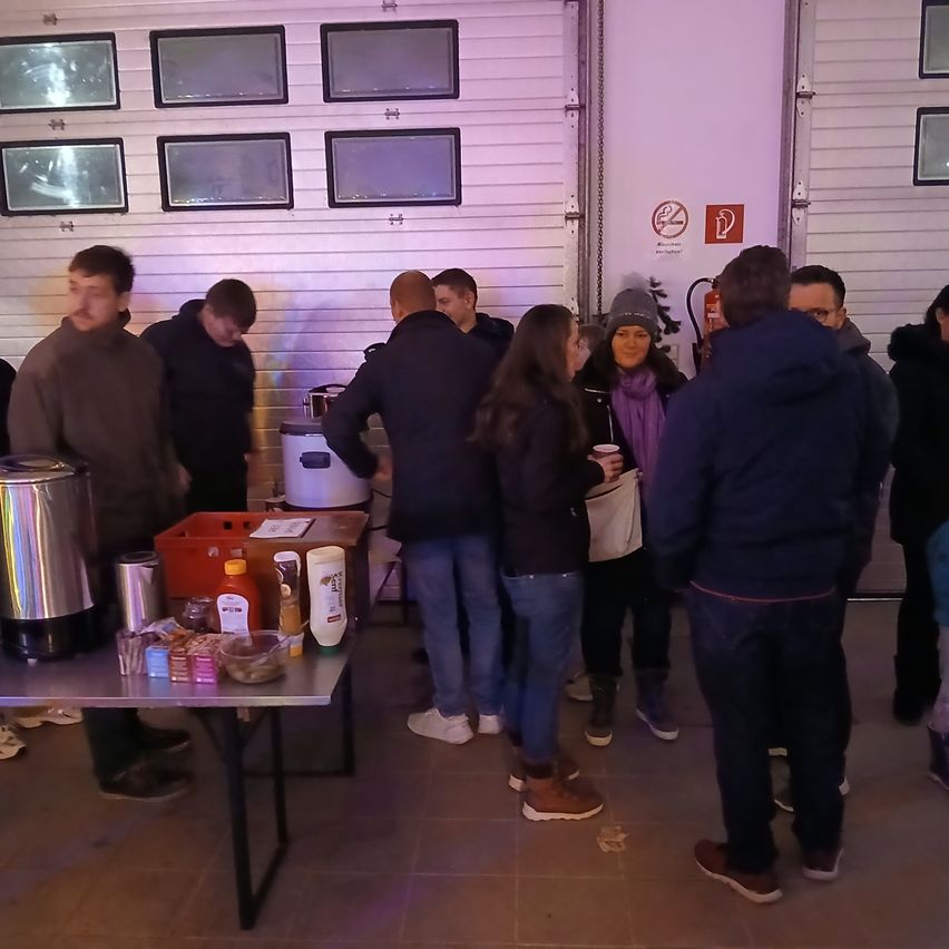 Eine Gruppe von Menschen versammelt sich um einen Tisch mit verschiedenen Gegenständen in einem Raum. Sie scheinen auf etwas zu warten. An der Wand hängen Bilderrahmen.