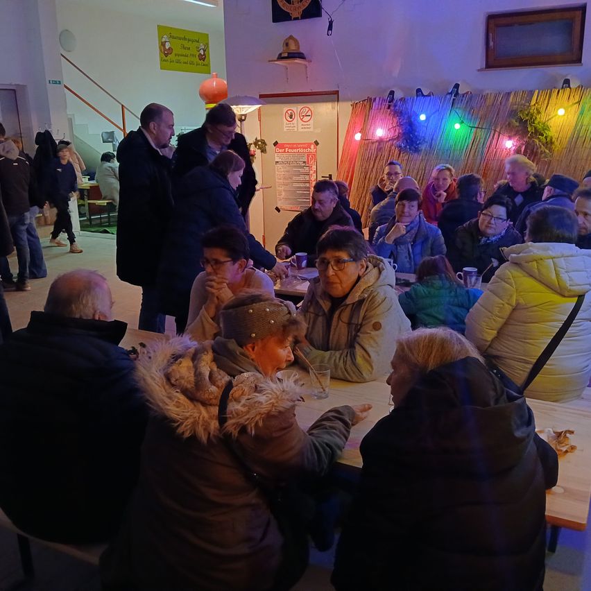 Mehrere Menschen sind in einem Raum versammelt, einige sitzend und andere stehend. Ein Tisch mit Tassen und eine Frau mit einer pelzgefütterten Jacke. Ein Rauchverbotsschild ist an der Wand, und es gibt bunte Lichter.