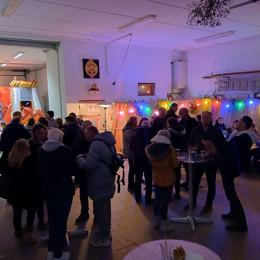 Ein großer Innenbereich ist mit vielen Menschen gefüllt, einige stehen und einige sitzen. Bunte Lichter schmücken den Raum. Ein Mann mit Rucksack steht neben einem Tisch mit Getränken.