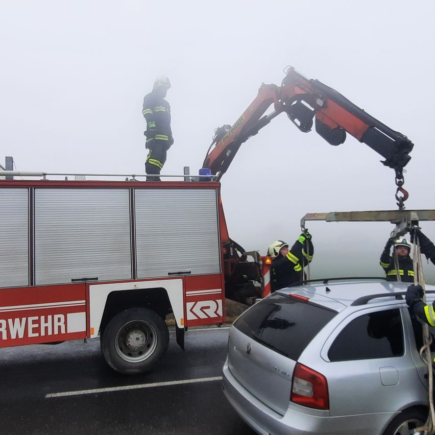 Feuerwehrleute stehen auf einem Feuerwehrauto und benutzen einen Kran, um ein Auto auf einer nebligen Straße anzuheben.