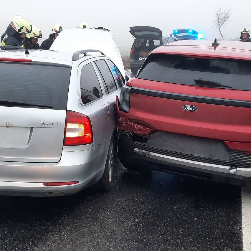 Rettungskräfte in Helmen inspizieren einen silbernen und einen roten Wagen mit Schäden auf einer nebligen Straße. Der rote Wagen hat ein Ford-Logo.