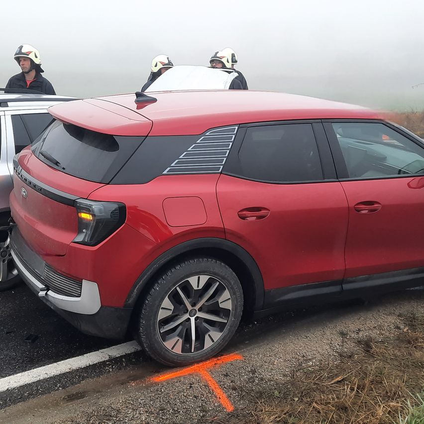 Ein roter SUV ist am Straßenrand geparkt, hinter ihm stehen drei Personen mit Helmen. Der SUV hat eine weiße Linie an der Stoßstange. Das Gebiet ist neblig.