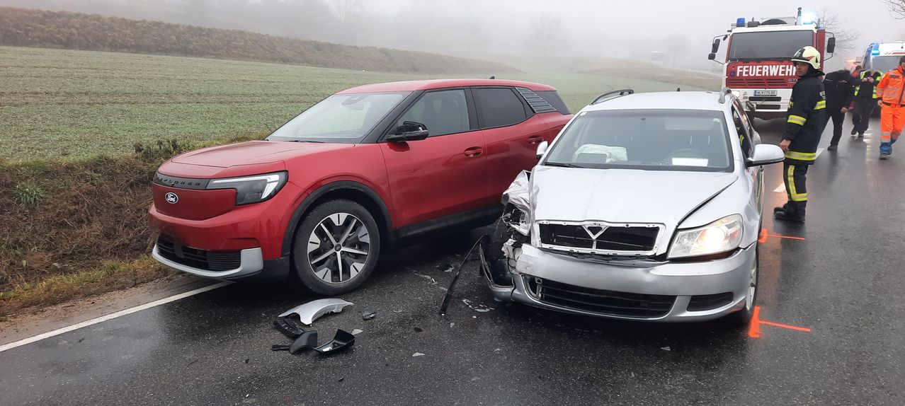 Zwei Autos sind auf einer nebligen Straße geparkt. Das rote Auto steht links, das silberne rechts. Beide Autos haben Schäden an der Front.