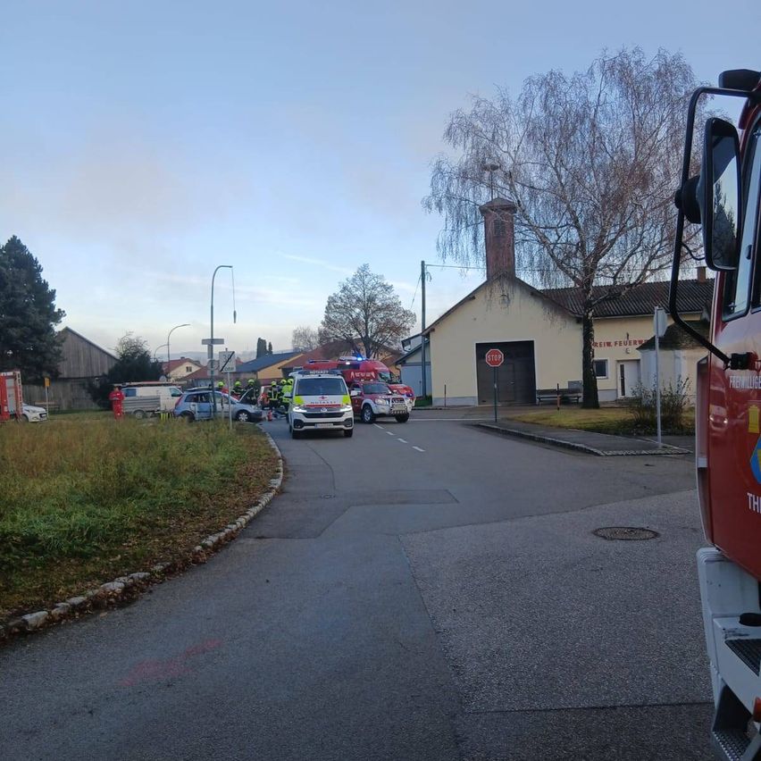 Einsatzfahrzeuge stehen auf einer Straße mit Gebäuden und Bäumen im Hintergrund. Eine Person in einem roten Anzug steht in der Nähe der Fahrzeuge.