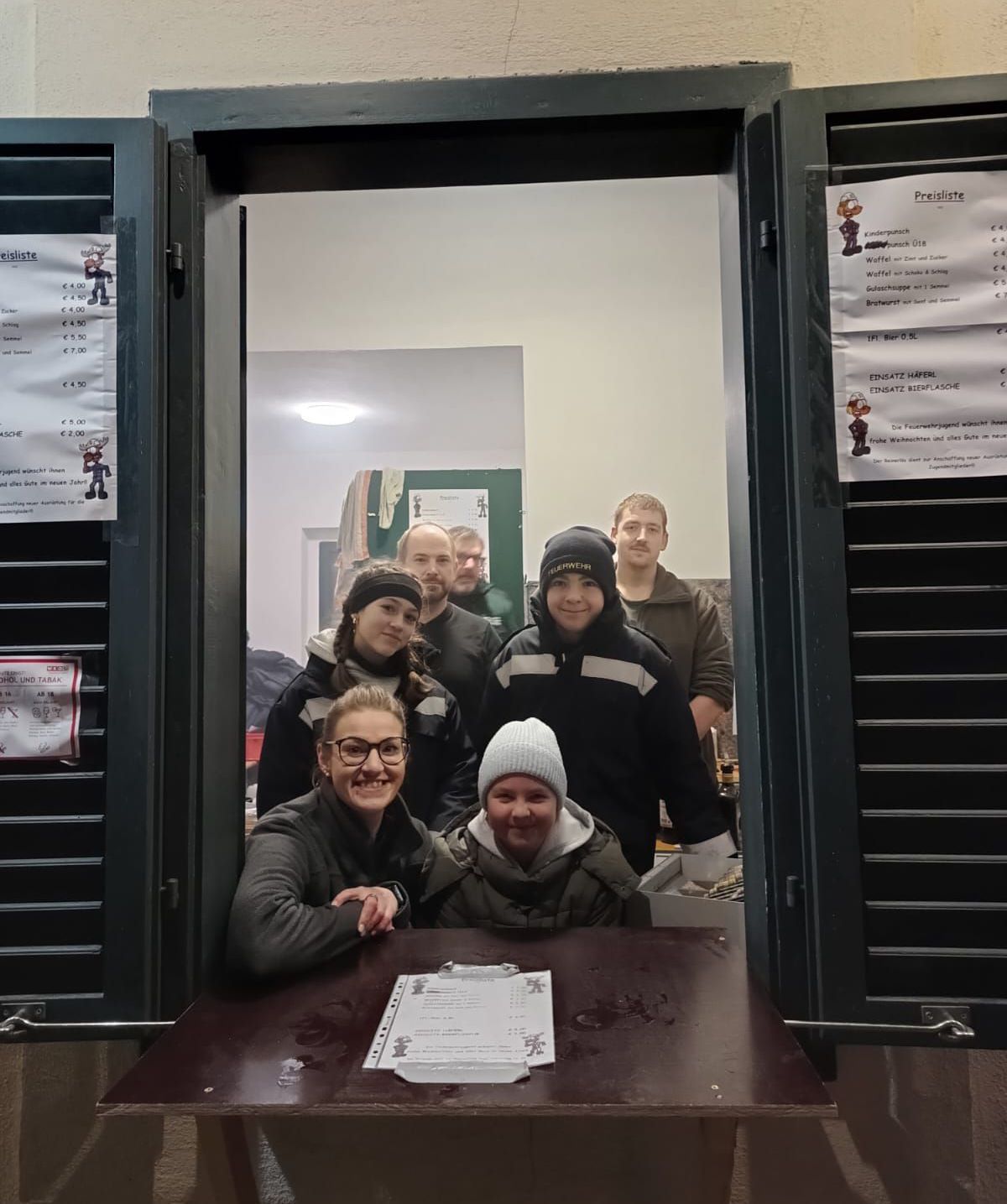 Eine Gruppe von Menschen in Winterkleidung steht vor einem Tresen. Sie lächeln für ein Foto. Papiere liegen auf dem Tresen. Poster und ein Schild sind an der Wand angebracht.