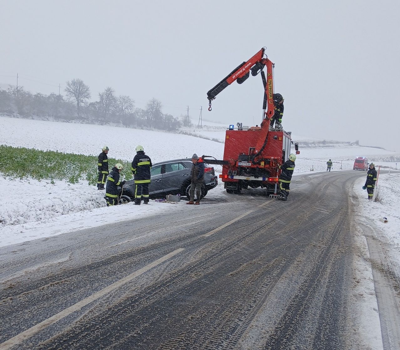 Ein Rettungsteam kümmert sich auf einer verschneiten Straße um einen Verkehrsunfall, ein Feuerwehrauto und Personal sind vor Ort.