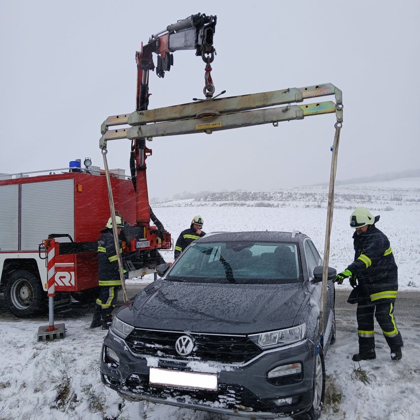 Feuerwehrleute verwenden einen Kran, um ein graues Volkswagen-Auto zu heben, das im Schnee feststeckt. Das Auto ist auf der verschneiten Straße positioniert, mit der Front zum Kameramann gerichtet.