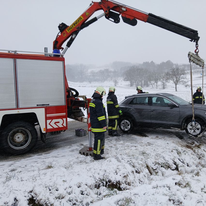 Feuerwehrleute in gelben und schwarzen Uniformen heben mit einem Kran ein Auto auf einer verschneiten Straße an.