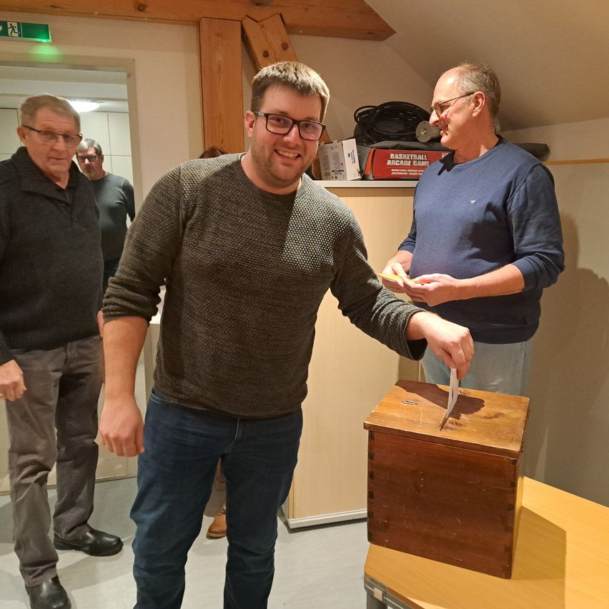 A man casts his vote into a wooden ballot box while two older men look on, with one holding a piece of paper. The scene takes place indoors.