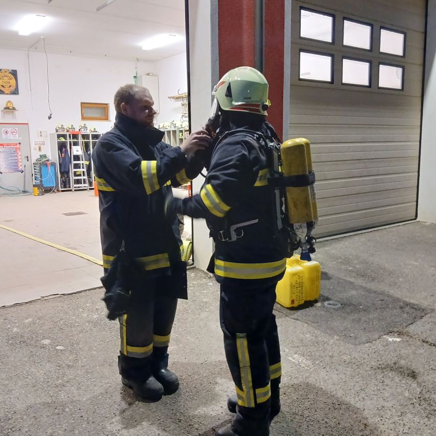 Two firefighters in full gear, one adjusting the other's helmet, standing in a garage with an open door.