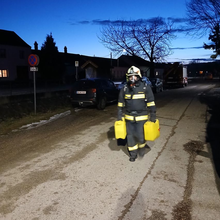 A firefighter wearing a protective suit walks down a street at night, carrying yellow containers. Behind him are parked cars and a building with a red sign.
