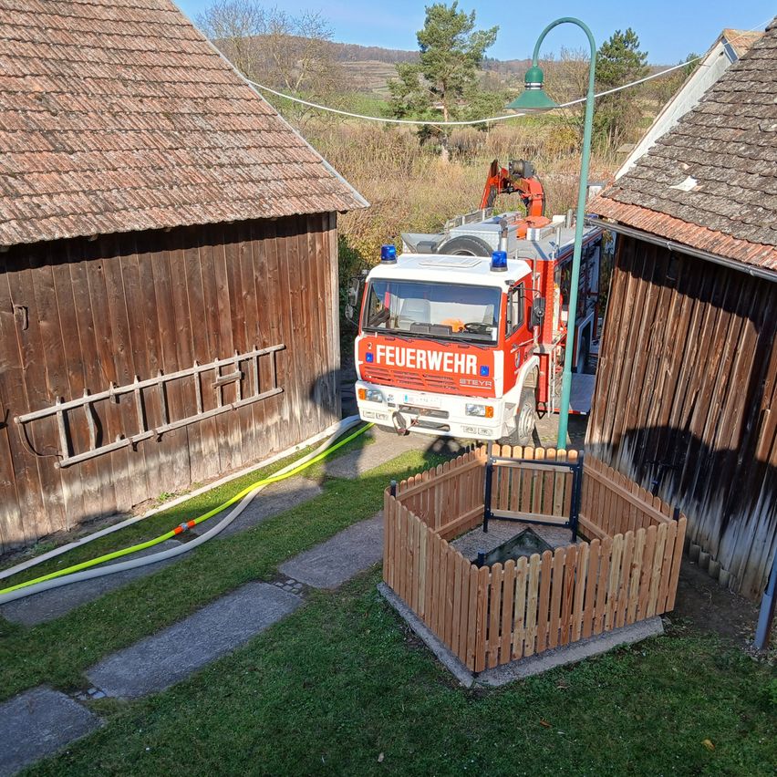 A fire truck is parked near a wooden structure in a rural area. A green lamp post is standing nearby. The area is surrounded by grassy lawns.