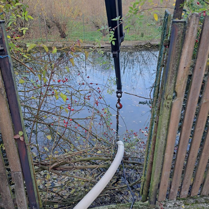 A view of a murky pond with a white pipe extending into the water. A crane with a hook is positioned above the pond, near a wooden fence.