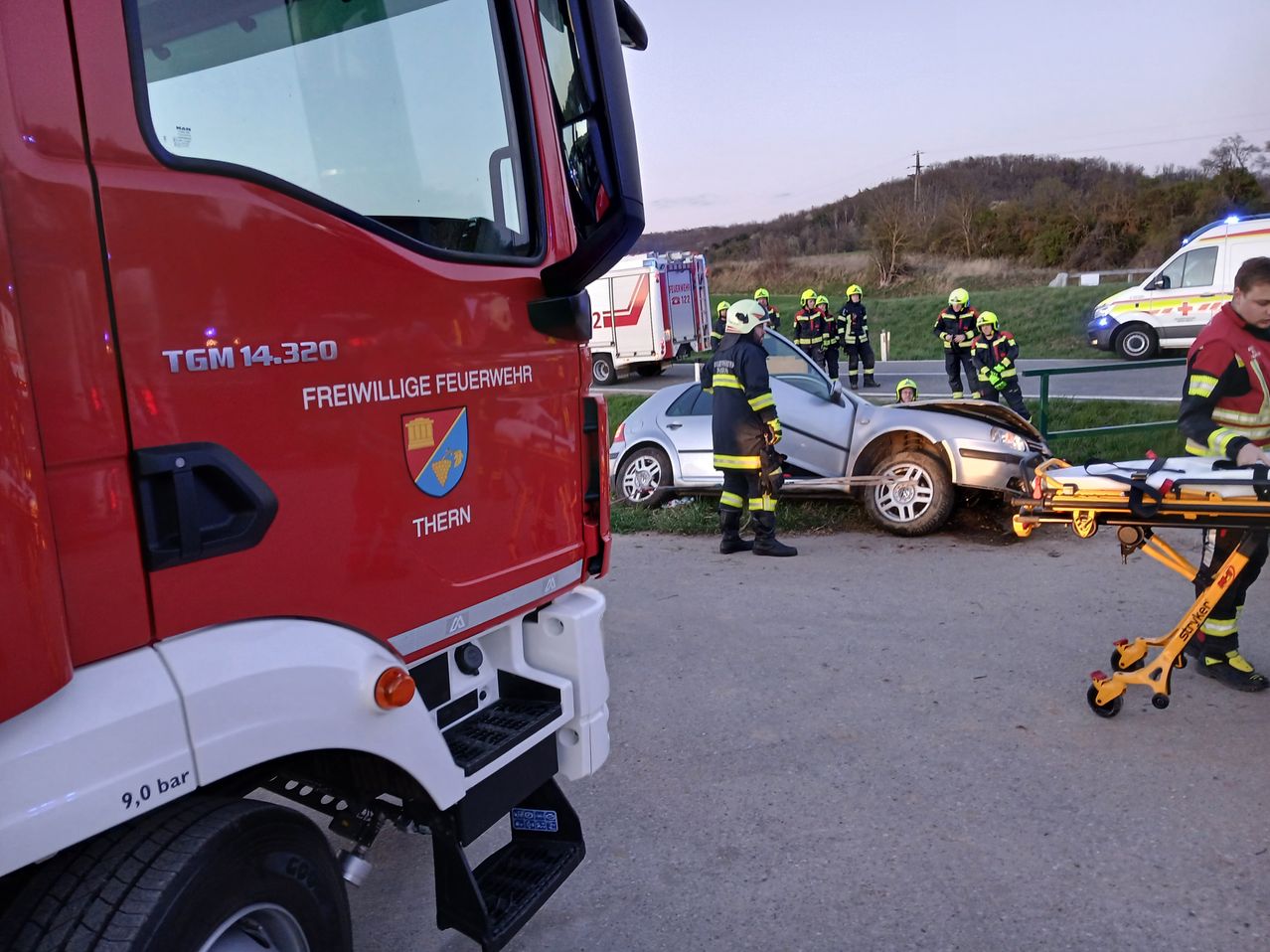 Ein freiwilliger Feuerwehrmann inspiziert ein beschädigtes Auto am Straßenrand, in der Nähe eines Feuerwehrwagens.