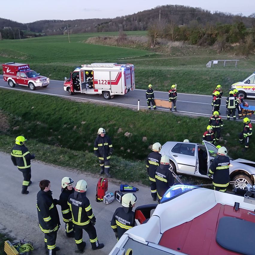 Rettungskräfte und Fahrzeuge sind am Unfallort eines Autounfalls. Das Feuerwehrfahrzeug hat seine Tür geöffnet und Menschen kümmern sich um eine verletzte Person.