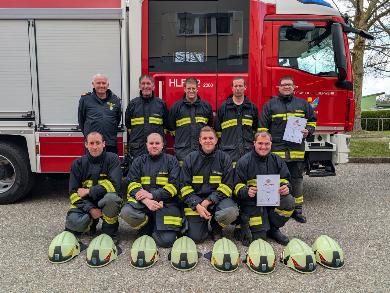 Eine Gruppe Feuerwehrleute in Uniform posiert mit Helmen und Zertifikaten vor einem Feuerwehrwagen. Sie lächeln für ein Foto.