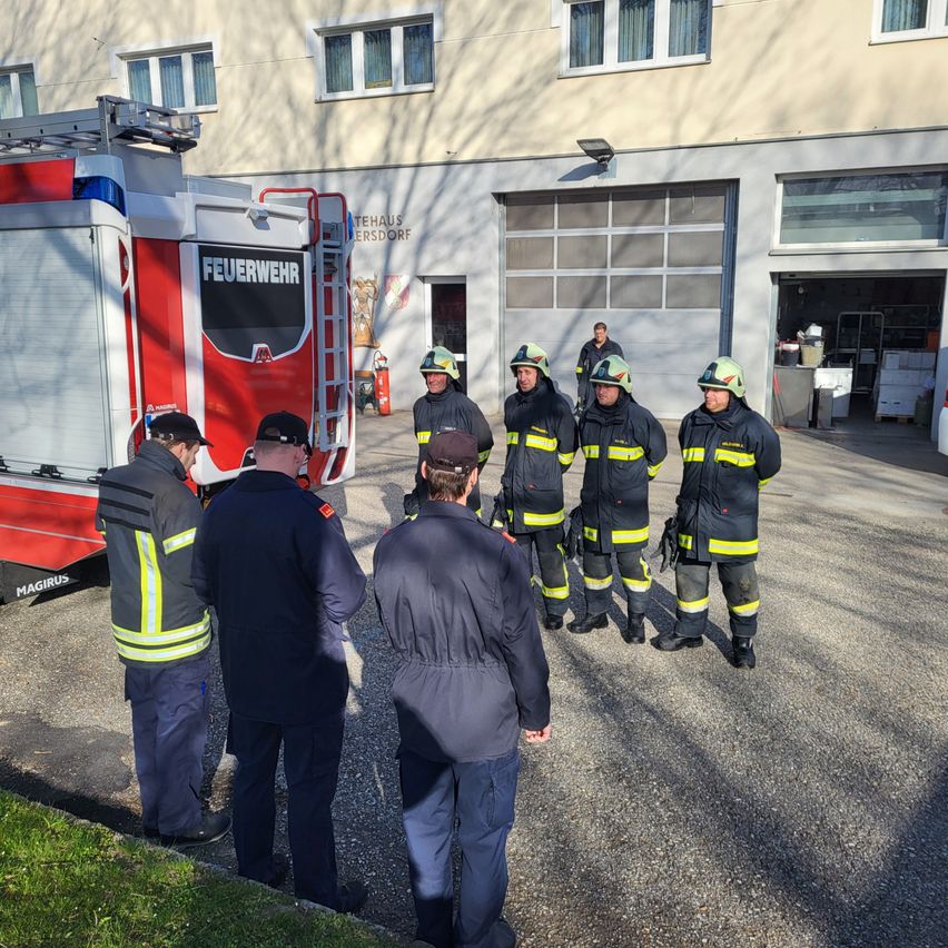 Eine Gruppe von Feuerwehrleuten in Uniformen versammelt sich vor einer Feuerwache, möglicherweise im Gespräch. Ein Feuerwehrauto ist neben ihnen geparkt.