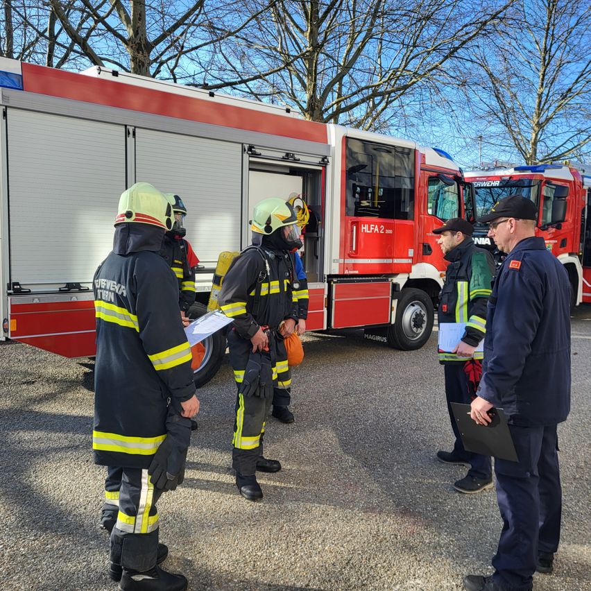 Feuerwehrleute in Schutzkleidung stehen vor einem roten Feuerwehrauto. Einer von ihnen hält ein Klemmbrett. Sie besprechen etwas. Das Auto hat ein Nummernschild, das 'HLAF 2' liest.