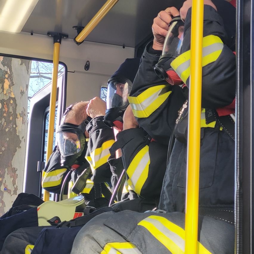 Drei Feuerwehrleute sitzen in einem Bus und tragen Schutzausrüstung und Masken. Einer hat einen Helm neben sich.