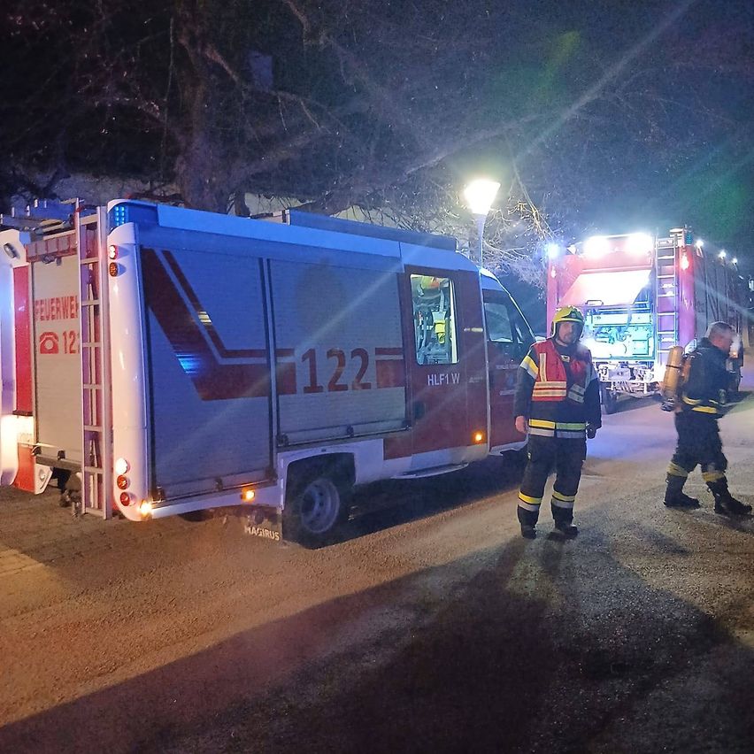 Two firefighters stand on the street at night with emergency vehicles parked. One has a helmet and a mask. The other holds a fire extinguisher.