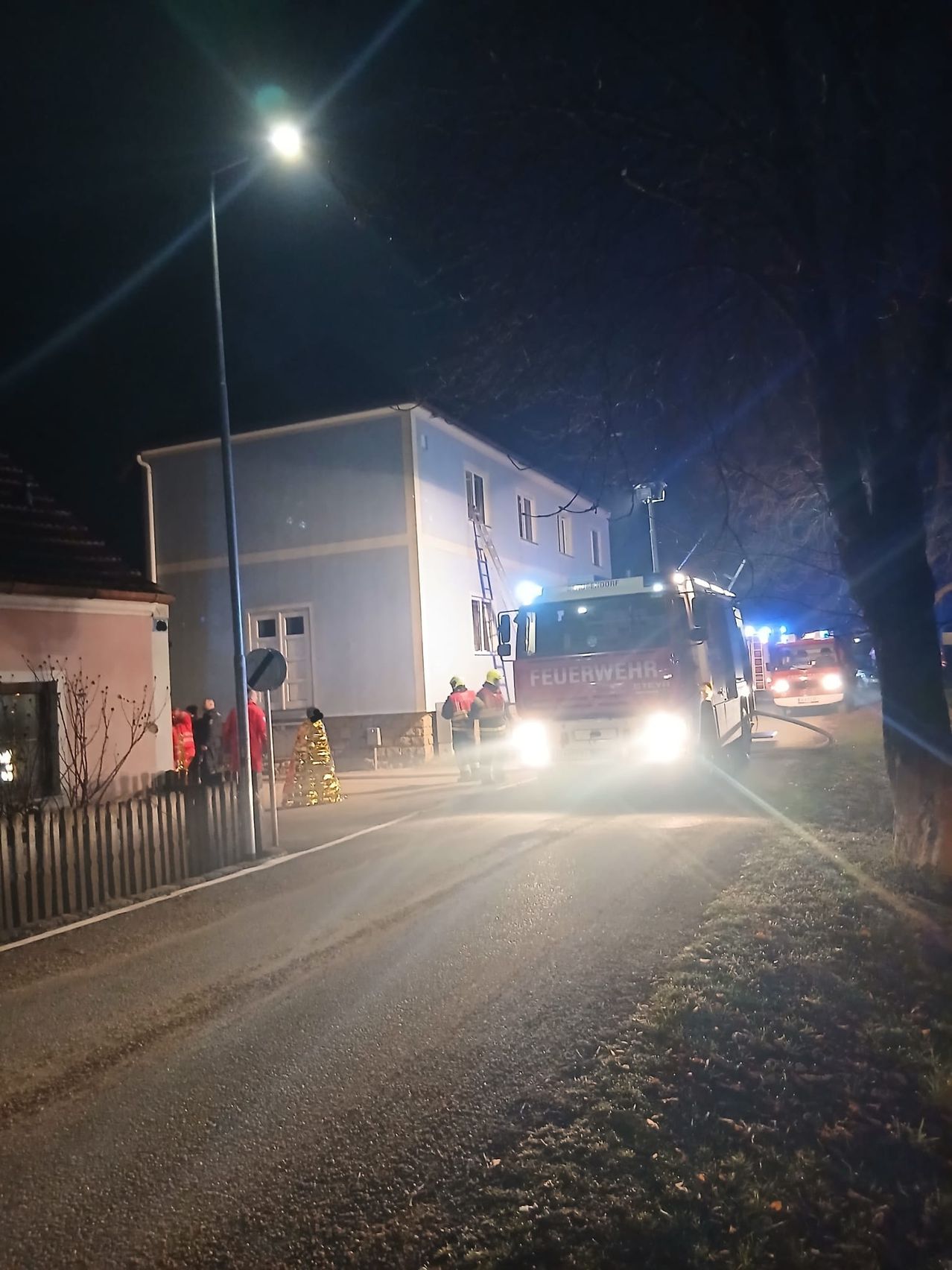 At night, a fire truck is parked on the street with firefighters attending to a building on the left. A ladder is leaned against the building, and a fire hydrant is connected to a hose.