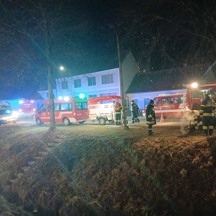 Emergency vehicles are parked in front of a building at night. Several firefighters are present, some standing and others near the vehicles. Trees and a streetlight are in the foreground.