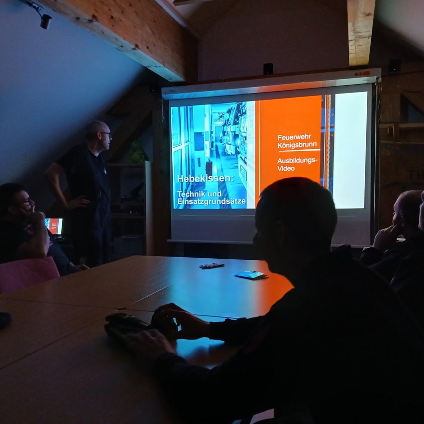 Several people are seated in a room watching a presentation. A man in a black shirt stands near a screen. A projector screen shows a slide with text and a video.