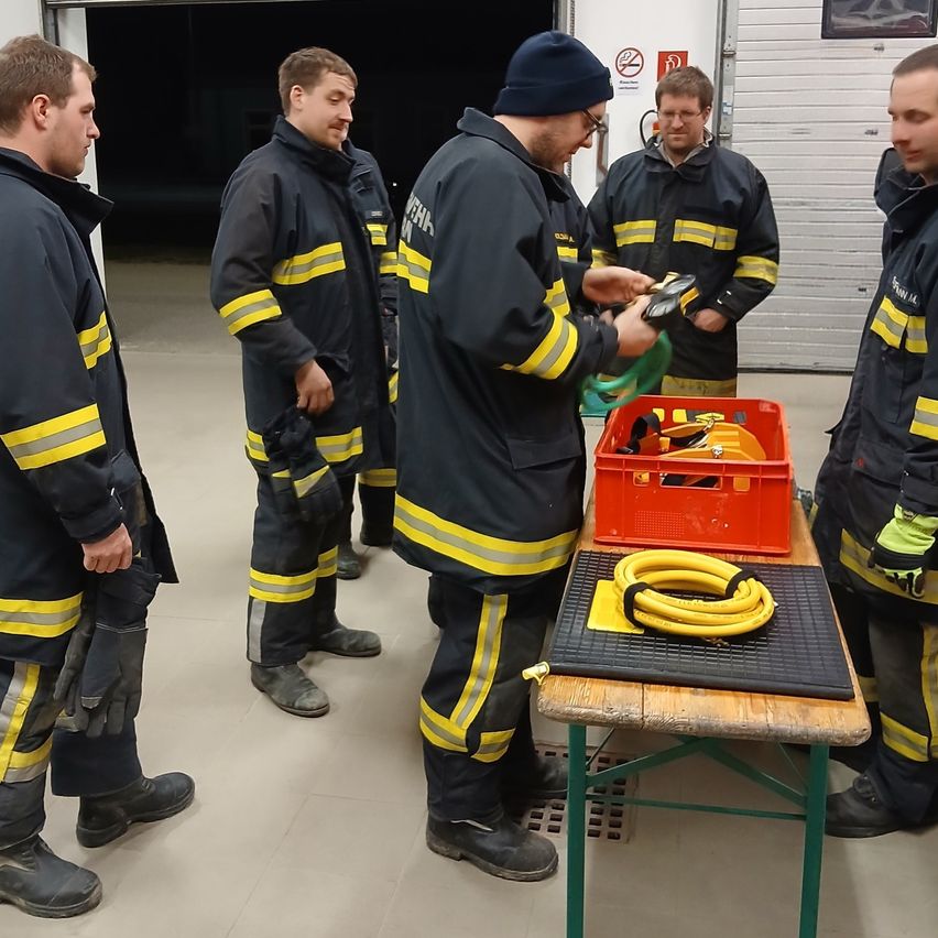 A group of firefighters are standing around a table with tools. One is demonstrating something while others watch. They wear uniforms with yellow stripes.