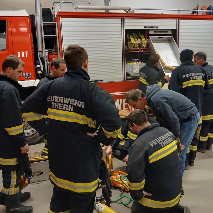 A group of firefighters are gathered around a fire truck, some inspecting equipment. The fire truck is marked with 'SRF' and 'Feuerwehr Thern'.