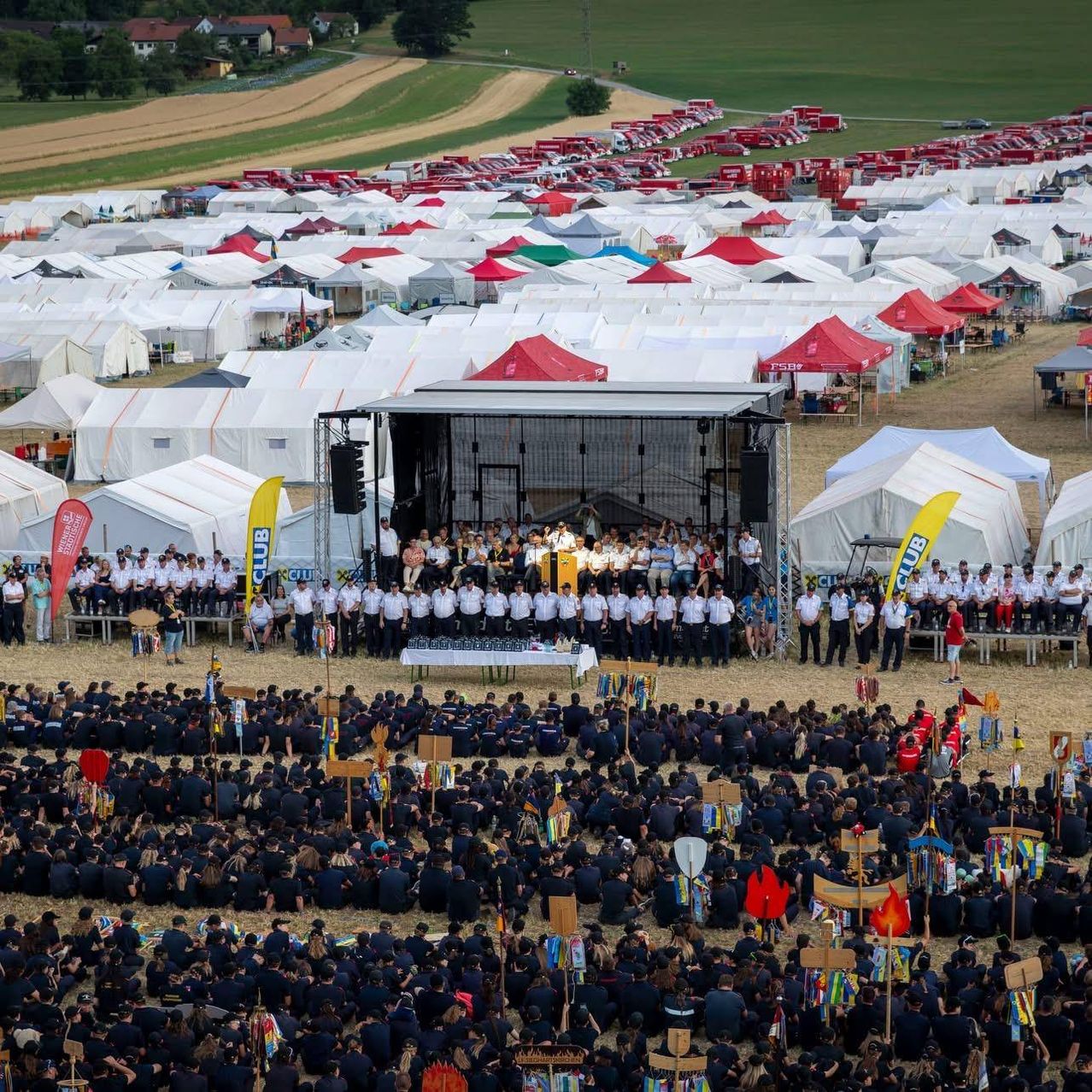 A large gathering of people in uniform, possibly a military event, is taking place in a field with many tents and a stage in the background.