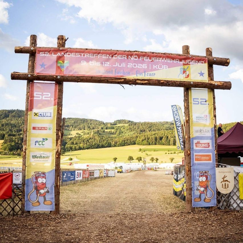 A wooden archway with banners advertising the 52nd Landestreffen der Nö Feuerwehrjugend event, held from July 9th to 12th, 2026. The backdrop features a scenic landscape with hills and greenery.