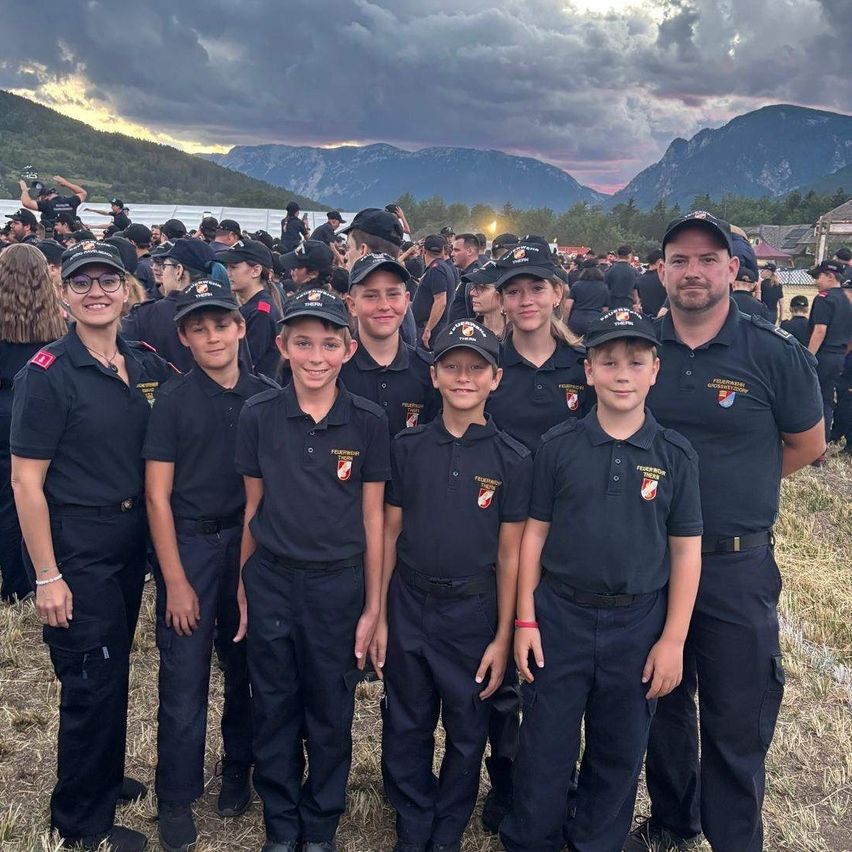 A group of young people in uniform, possibly firefighters, stand together in a field with mountains in the background.