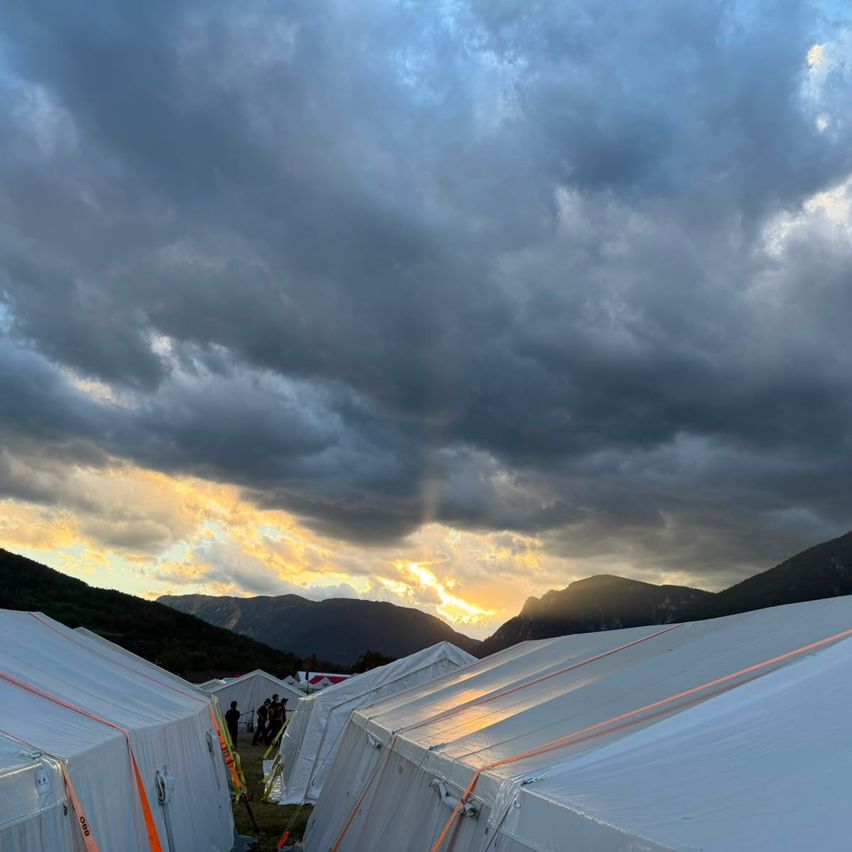 A row of white tents set up on a grassy field under a cloudy sky with a sunset in the background.