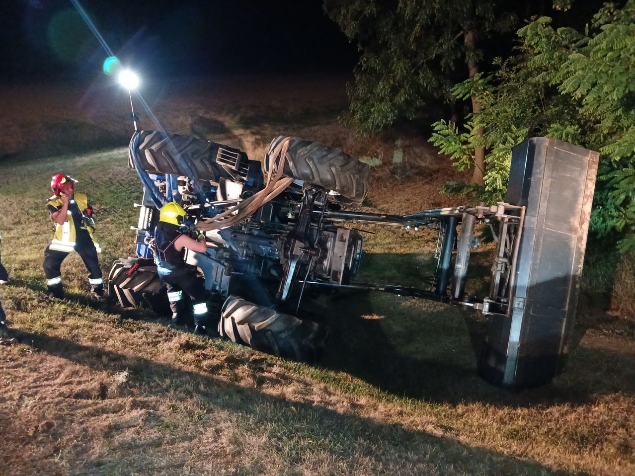 Ein Traktor ist nachts auf einem Grasfeld umgekippt, eine Person mit gelbem Helm arbeitet daran.