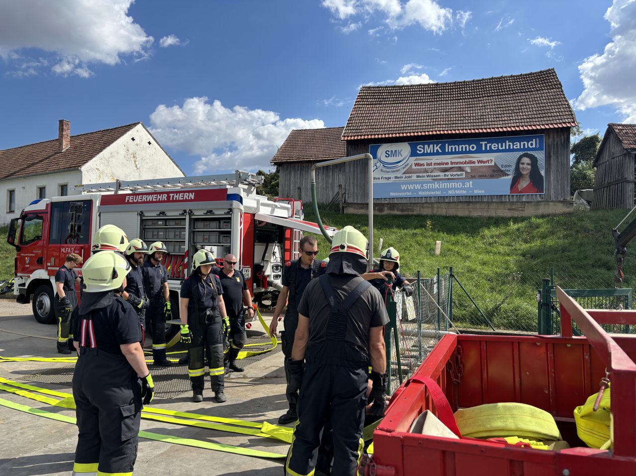 Eine Gruppe Feuerwehrleute in voller Ausrüstung steht vor einem Feuerwehrauto, mit einem roten Laderaum im Vordergrund und einem Haus mit einem Schild im Hintergrund.