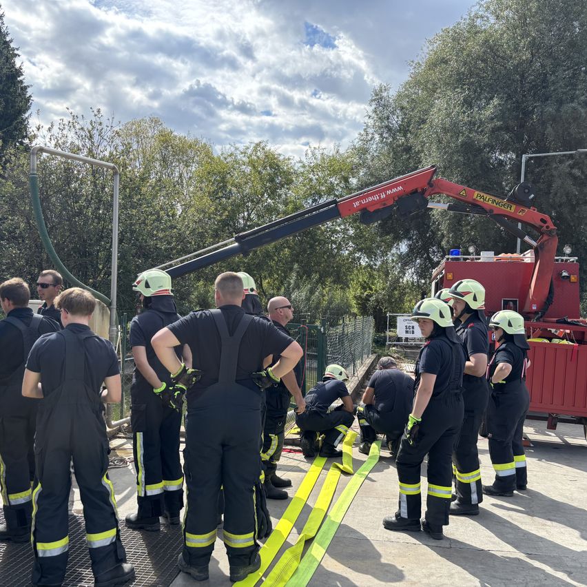 Eine Gruppe von Feuerwehrleuten in voller Ausrüstung, die um einen roten Kran herumstehen, wobei einige eine Aufgabe am Boden erledigen.