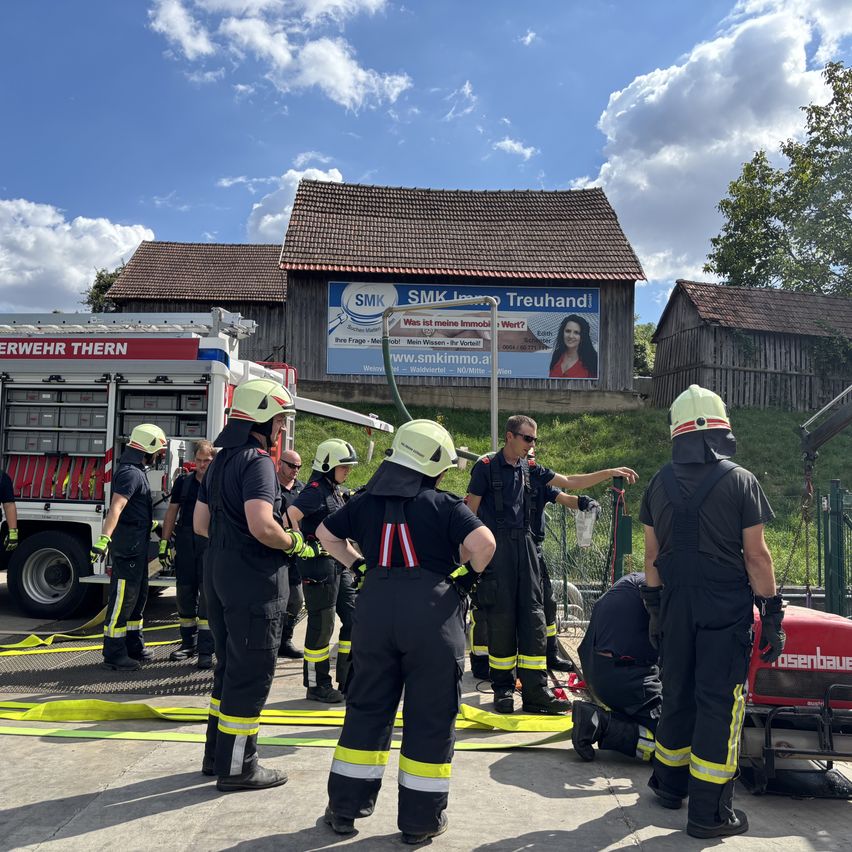 Eine Gruppe von Feuerwehrleuten in Uniform steht vor einem Haus mit einem Schild, um einen Feuerwehrwagen mit angeschlossener Schlauch herum.