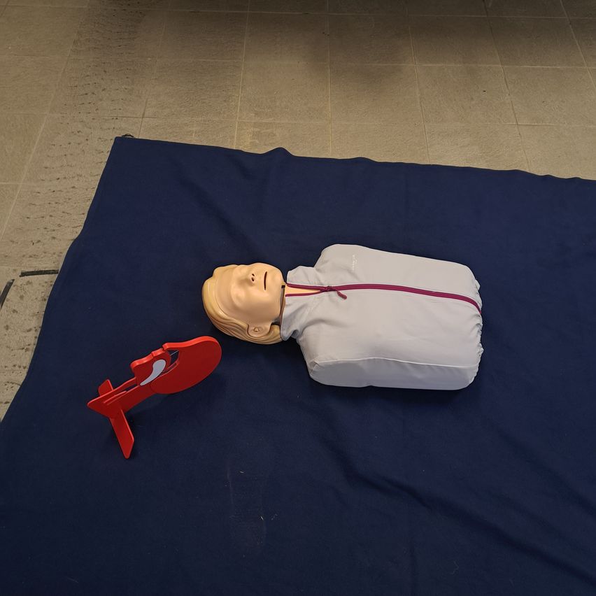 Eine CPR-Trainingspuppe liegt auf einer blauen Decke mit einem roten CPR-Leitfaden daneben auf einem gefliesten Boden.