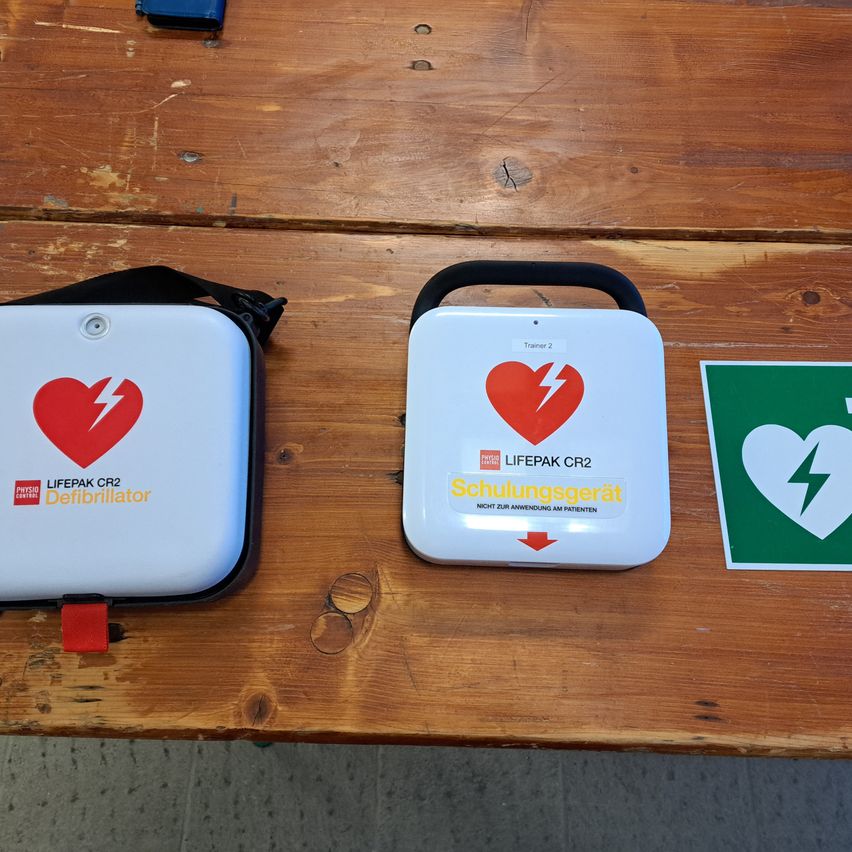 Zwei weiße Defibrillatoren mit roten Herzen und Blitzen sind auf einem Holztisch platziert. Der linke hat die Aufschrift 'LIFEPAK CR2 Defibrillator'. Der rechte hat die Aufschrift 'Schulungsgerat' und einen roten Pfeil nach unten.