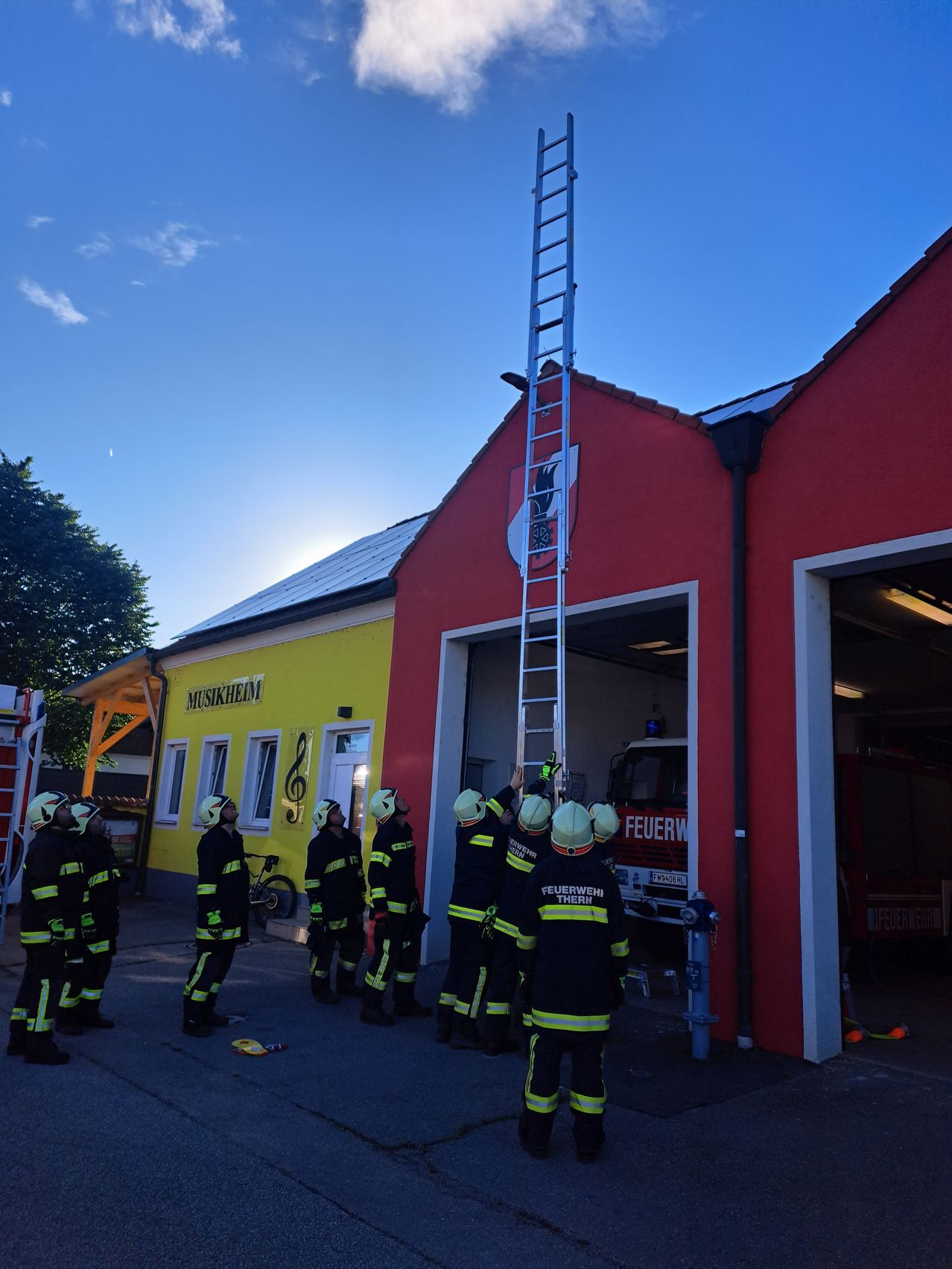 Eine Gruppe Feuerwehrleute in schwarzen Uniformen mit Helmen und Warnwesten stehen vor einem roten und gelben Gebäude, eine Leiter ist auf dem Dach.